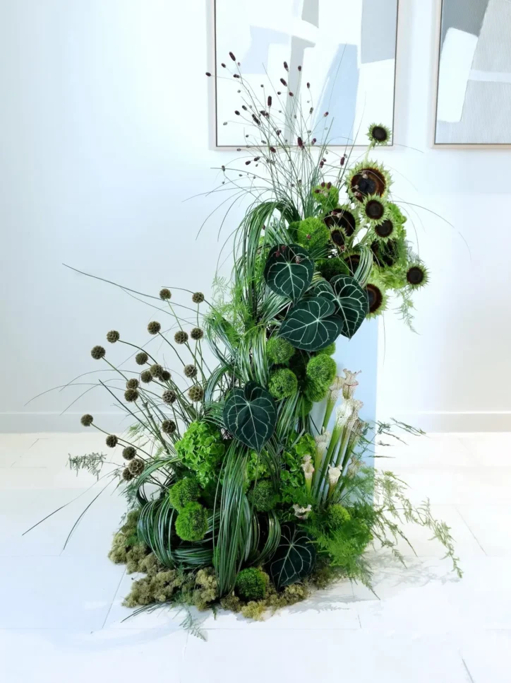 Tall green floral installation with dark-centered sunflowers, anthurium leaves, and ornamental grasses
