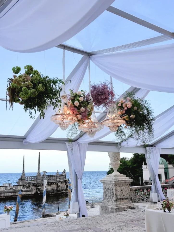 Floral and greenery arrangements hanging from crystal chandeliers under clear-top tent by the water