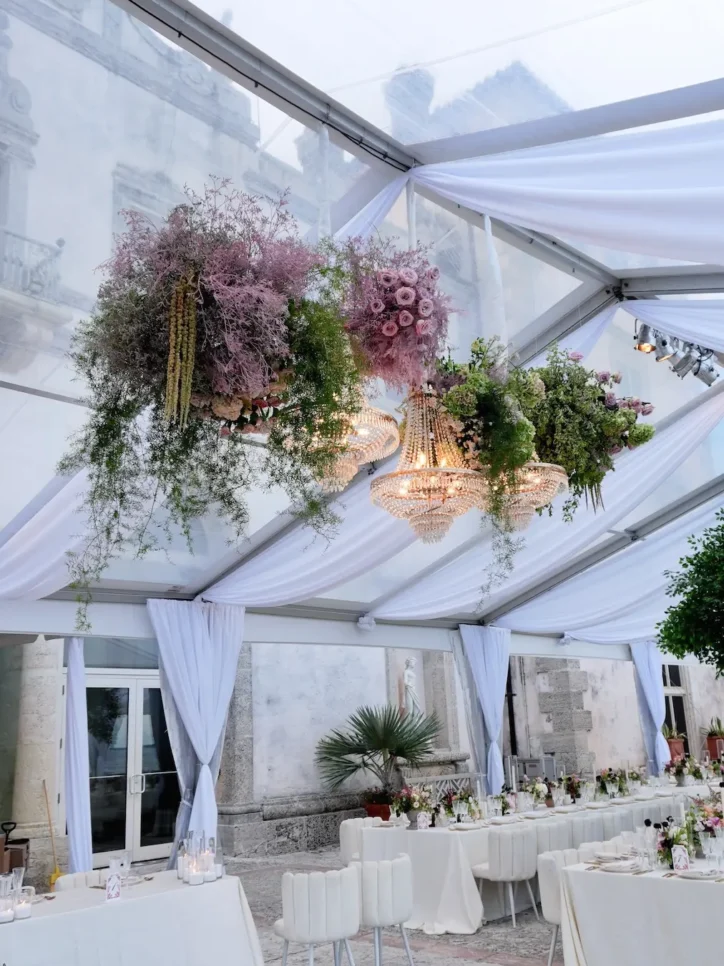 Pink floral chandelier installations above long reception tables under clear tent at historic estate