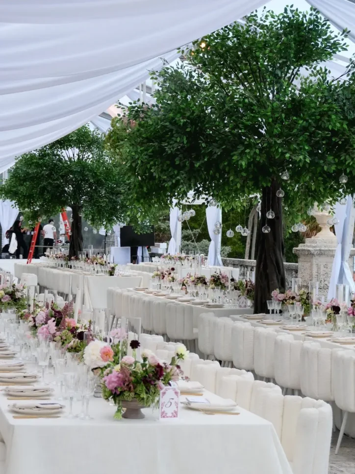 Pink and purple floral centerpieces on long reception tables under greenery trees and tented ceiling
