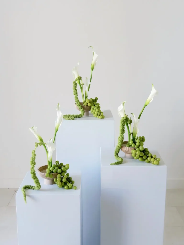 Three small calla lily and green grape arrangements with trailing amaranthus on white pedestals